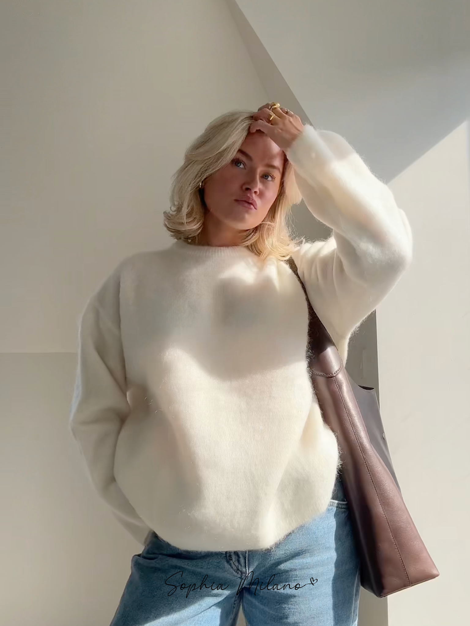 Blond kvinde i hvid sweater og blå jeans står i sollys, med hånden i håret. Bærer brun taske. Moderne mode, minimalistisk stil.