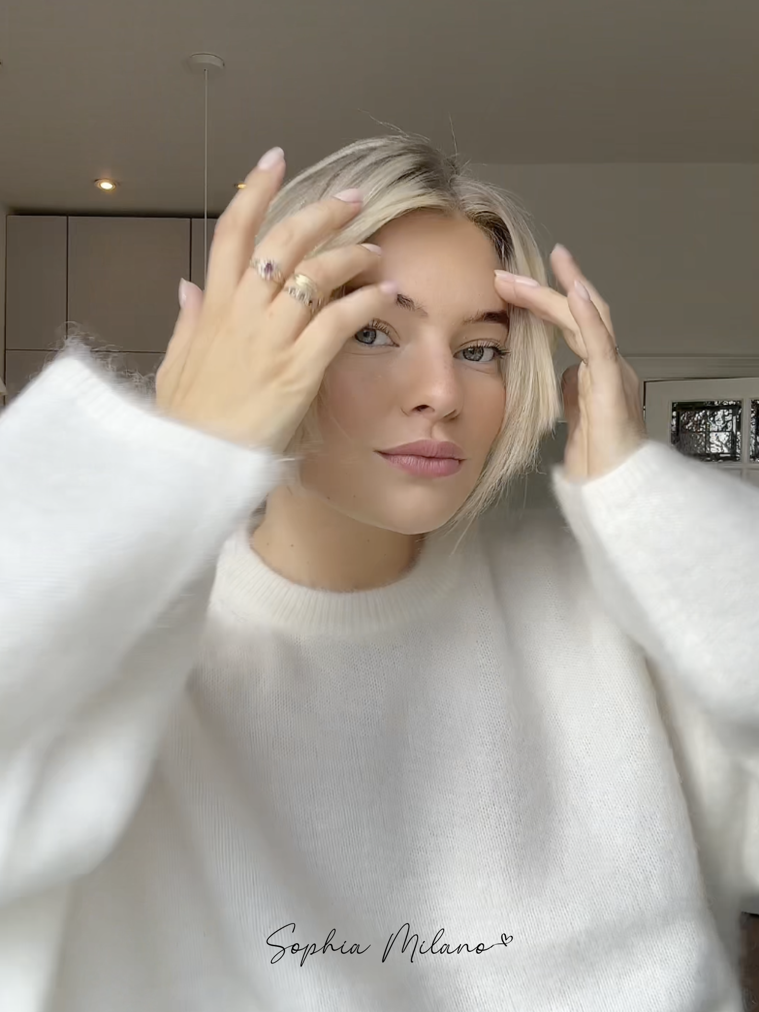 Ung kvinde med blondt hår iført hvid sweater, justerer sit hår i moderne interiør. Fokus på stil, mode og skønhed.