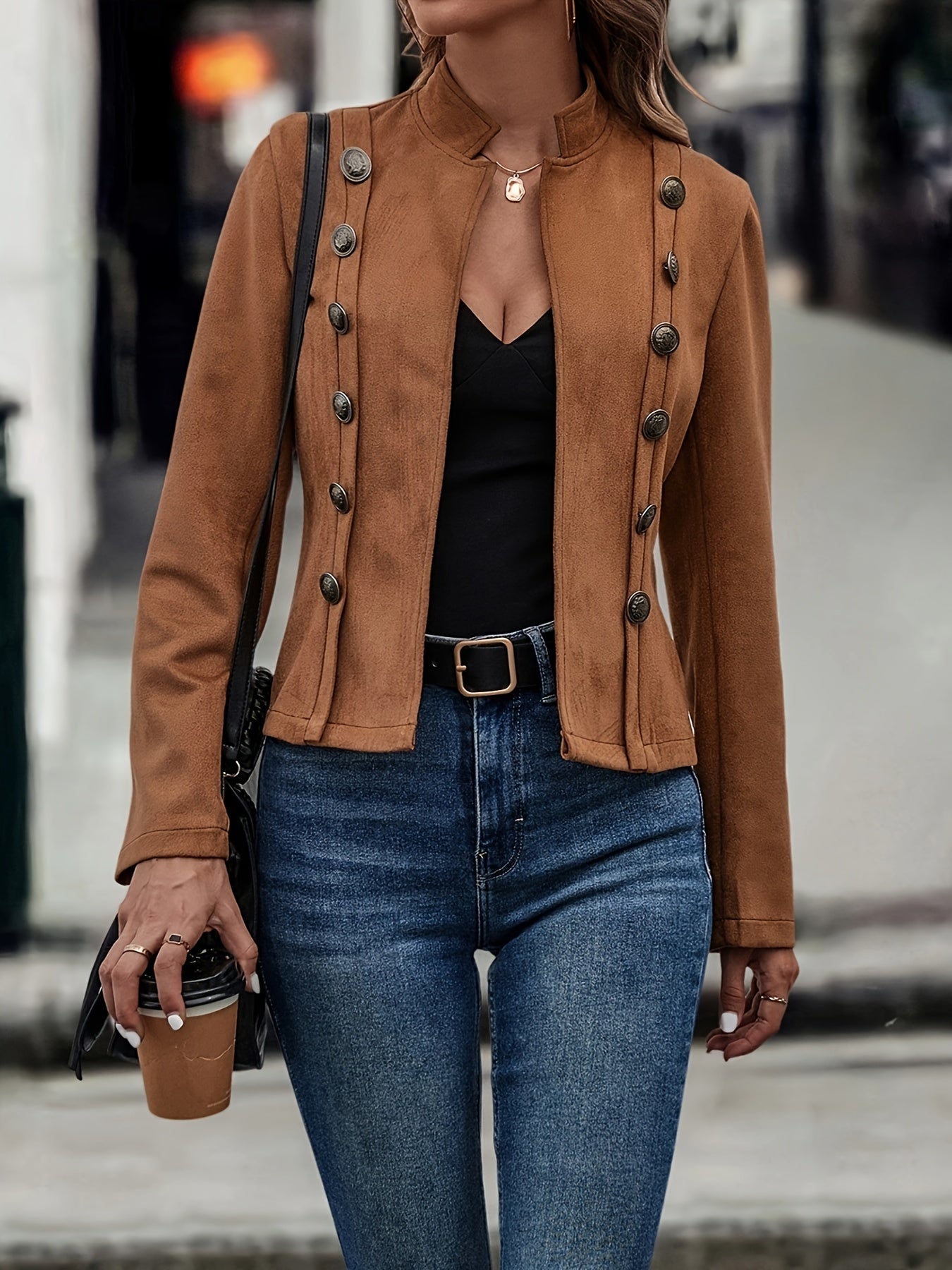 Kvinde iført brun jakke med knapper, sort top og jeans, holder en kaffekop. Mode, street style, efterårsjakke, casual outfit, byliv.