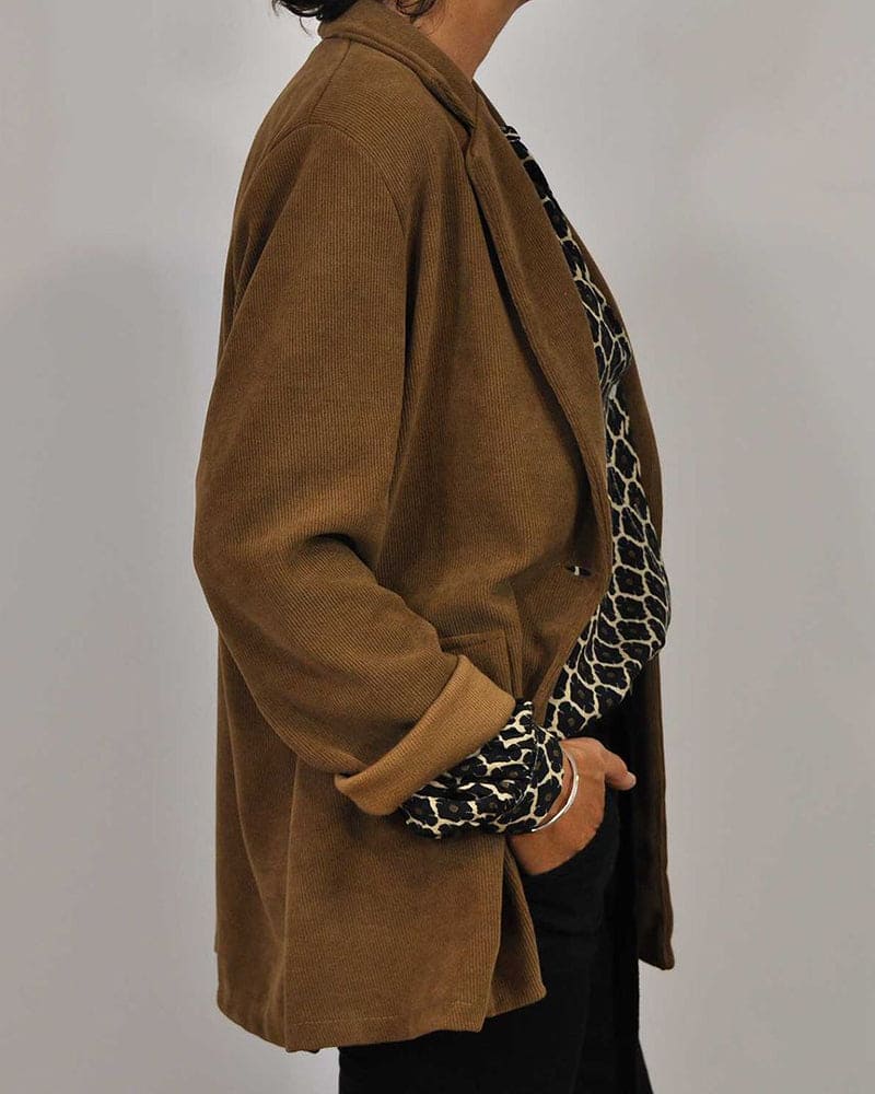 Kvinde iført brun blazer og mønstret bluse, set fra siden. Stilfuldt, moderne tøj til kvinder, perfekt til efterårsmode og kontorbrug.