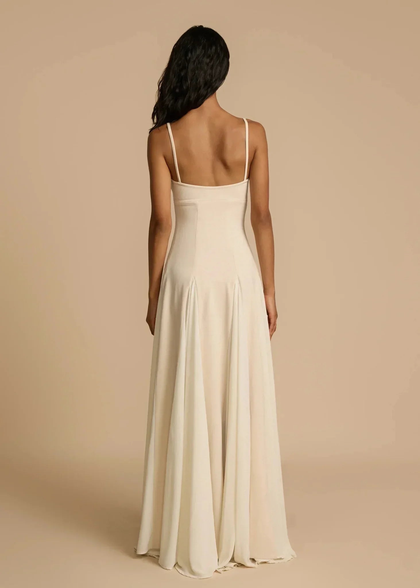 Kvinde i lang, elegant, beige aftenkjole med tynde stropper, set bagfra. Perfekt til formelle begivenheder. Stilfuldt og tidløst design.