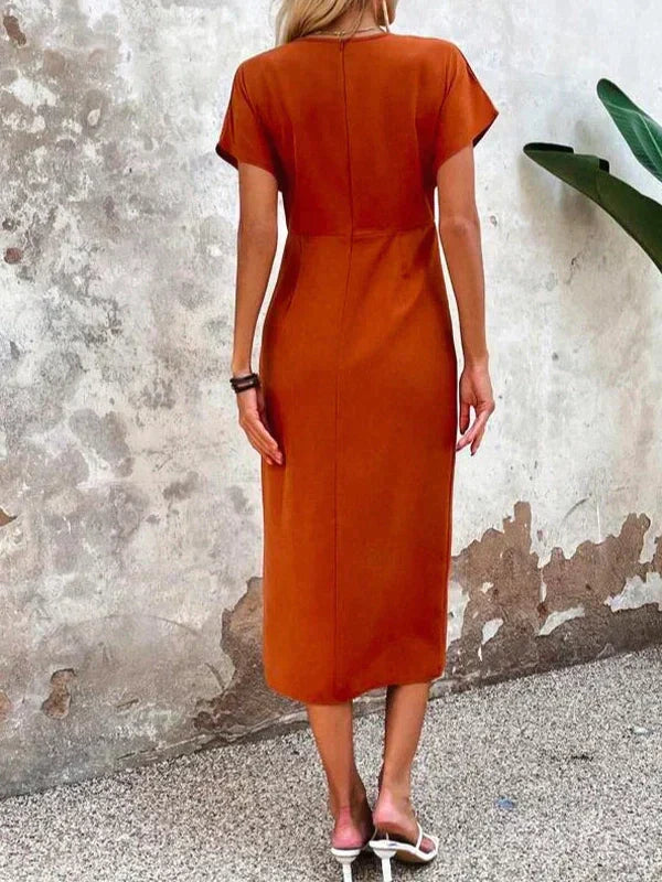 Kvinde i orange kjole set bagfra, står foran en rustik væg. Sommermode, elegant kjole, minimalistisk stil, udendørs baggrund.