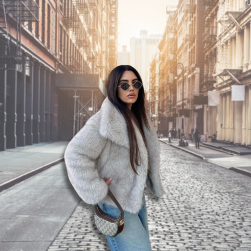 Kvinde i grå pelsjakke og solbriller på brostensbelagt gade i bymiljø. Mode, stilfuldt outfit, urbant landskab, trendy look.