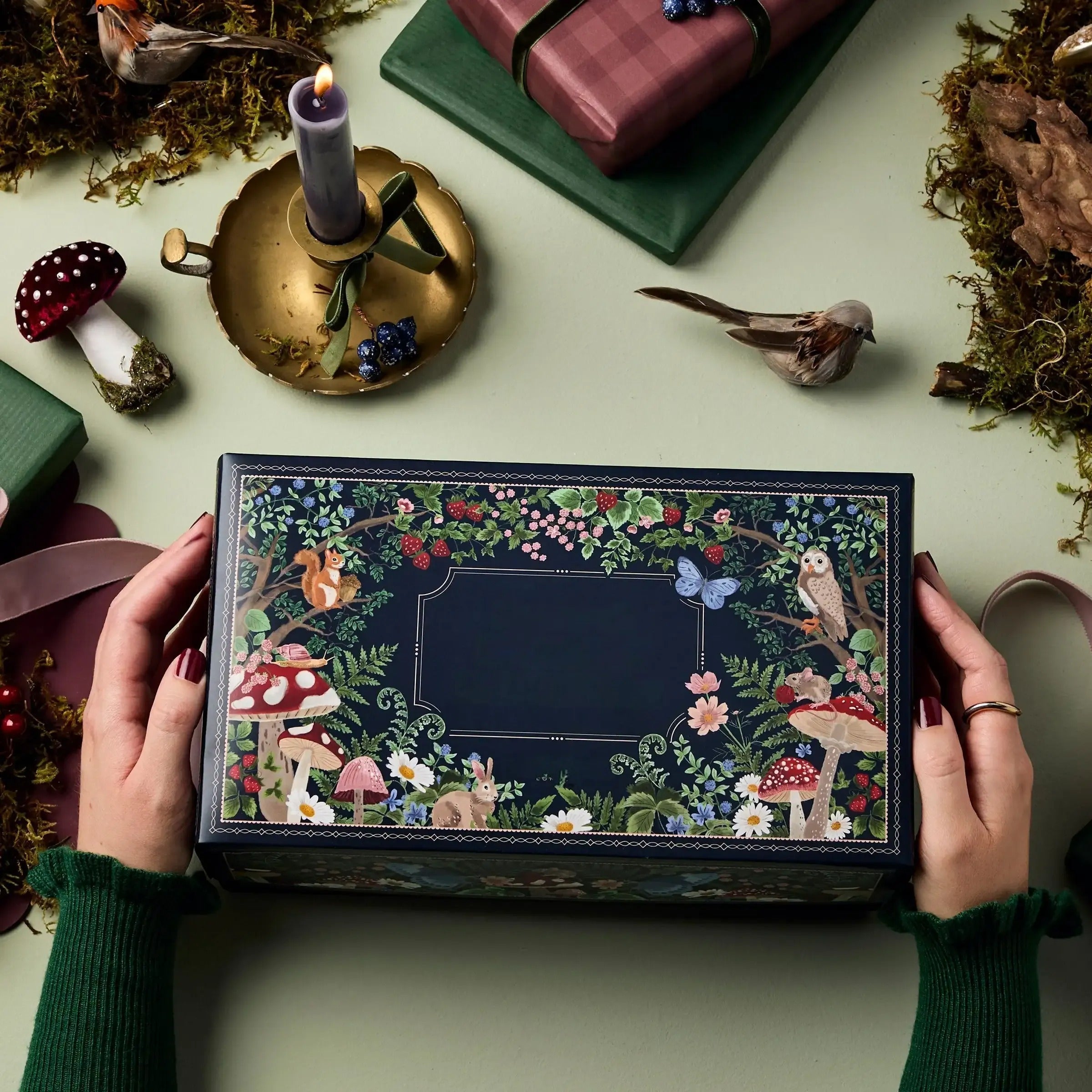 Hænder holder en dekoreret gaveæske med skovtema, omgivet af svampe, fugle og indpakkede gaver på et grønt bord. Perfekt til naturinspireret gaveindpakning.