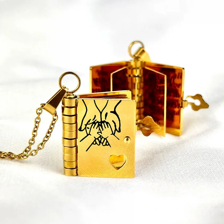 Guld bogformet vedhæng med hjerteudskæring og indgraveret design, åbent med synlige sider. Perfekt som romantisk smykke eller gaveidé.