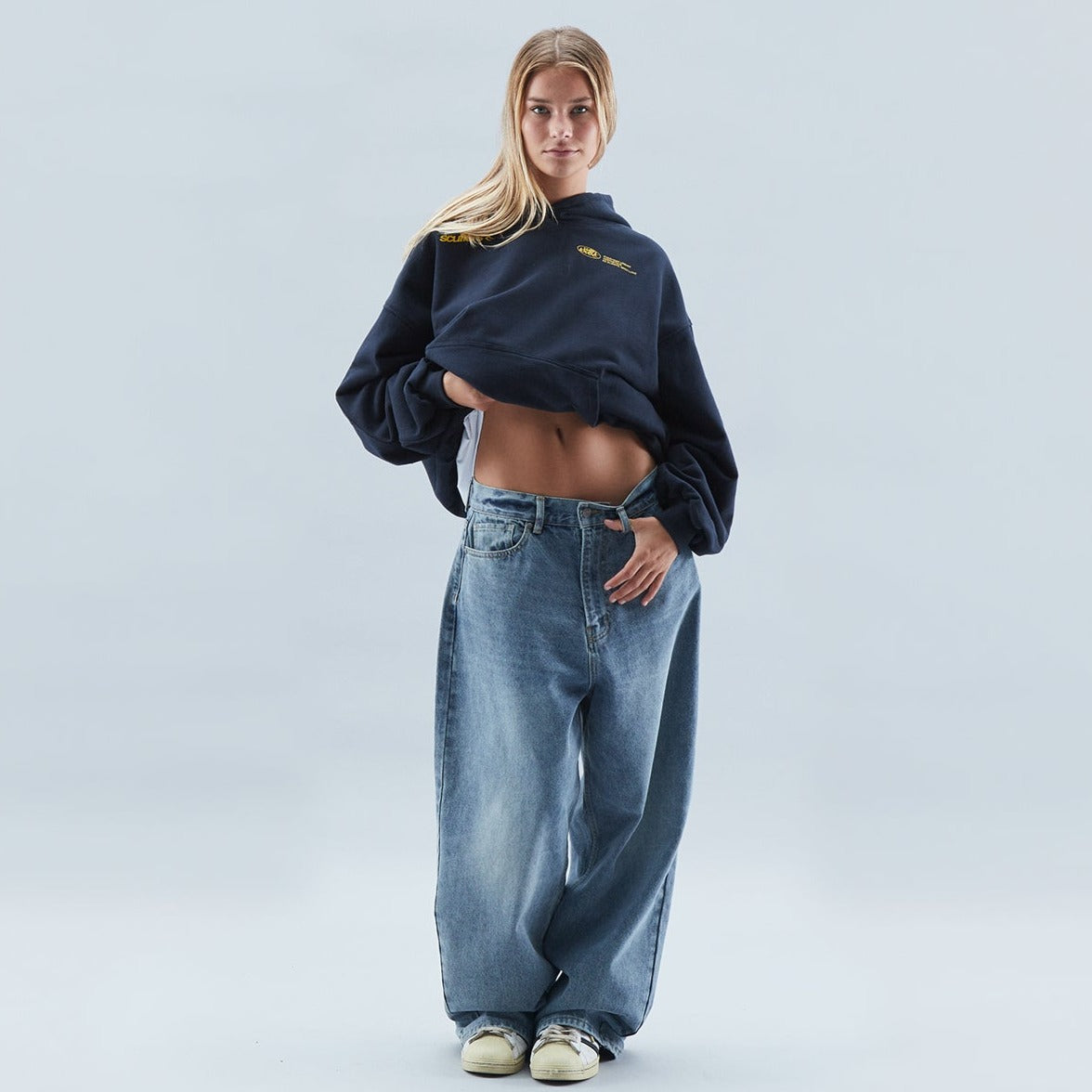 Ung kvinde i oversized jeans og mørk sweatshirt, viser mave. Moderne streetwear stil, afslappet look. Baggrund i lys grå.