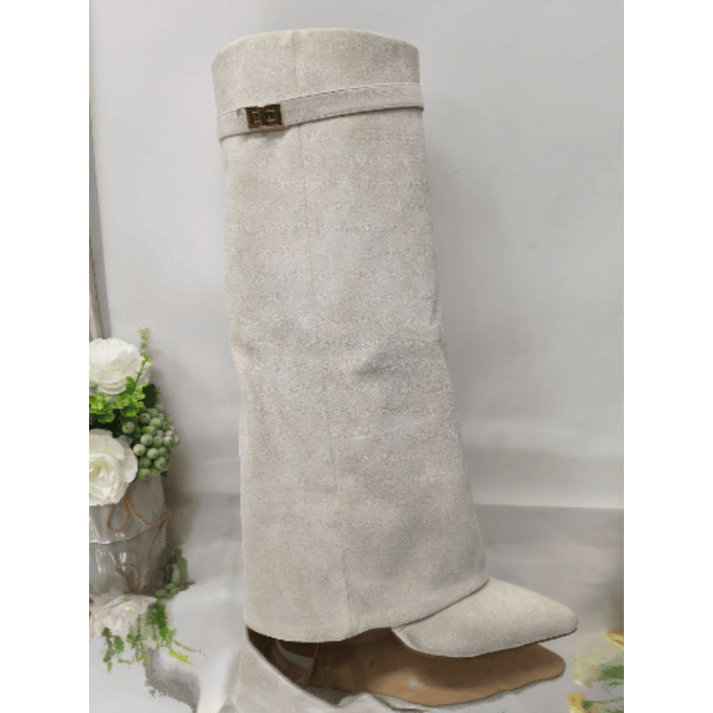 Hvid knæhøj støvle i ruskind med spids tå og dekorativt spænde, placeret på en skinnende overflade ved siden af hvide blomster.