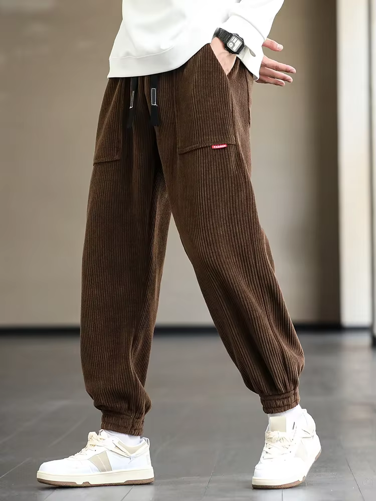 Brune fløjlsbukser til mænd, afslappet pasform, elastisk talje, moderne streetwear-stil, kombineret med hvide sneakers og hvid trøje.