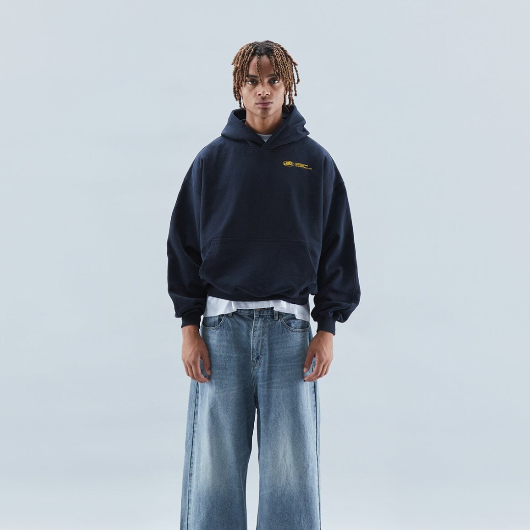 Ung mand i mørkeblå hættetrøje og brede jeans mod lys baggrund. Streetwear mode, afslappet stil, moderne ungdomstøj.