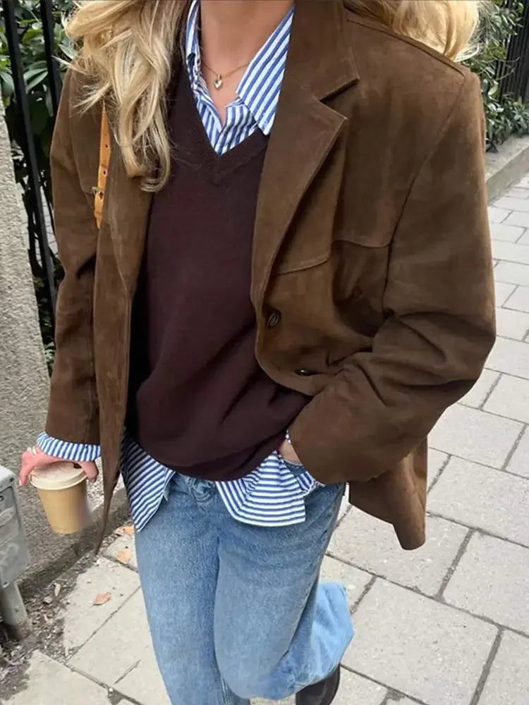 Kvinde iført brun ruskindsjakke, stribet skjorte og jeans, går på fortov med kaffekop i hånden. Mode, efterårstøj, casual stil.