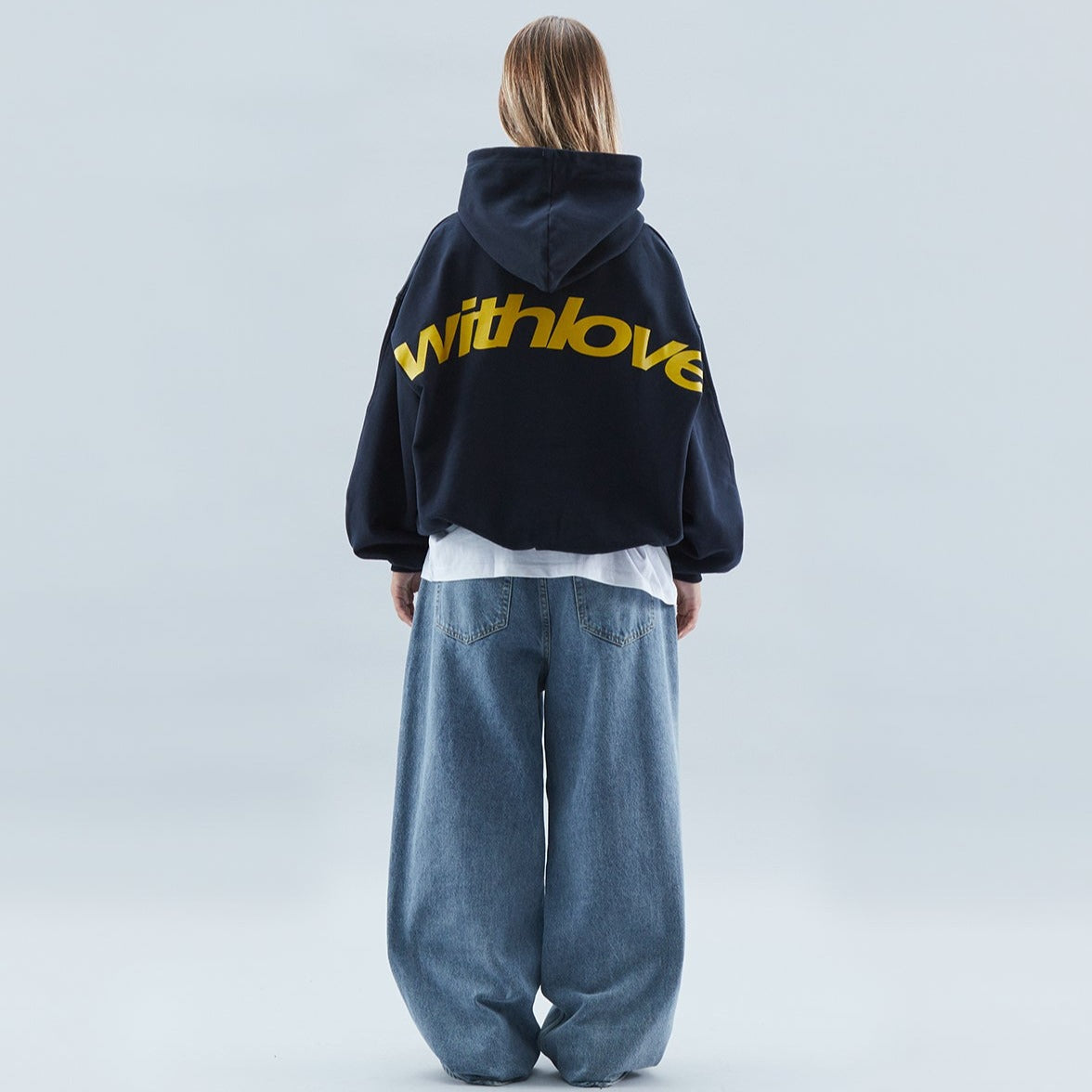 Kvinde i sort hættetrøje med gul "withlove" tekst, bagfra. Hun bærer løse, blå jeans. Moderne streetwear mode, minimalistisk baggrund.
