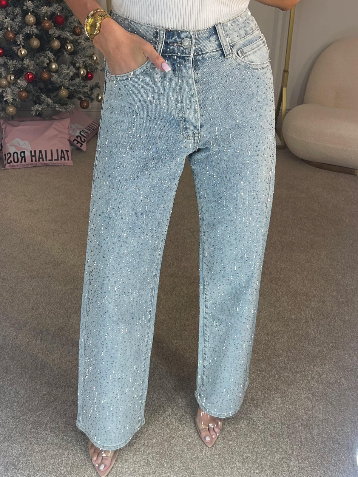 Kvinde iført lyseblå jeans med glitrende detaljer, stående på tæppe. Baggrund med juletræ og moderne stol. Mode, jeans, glimmer, tøjstil.