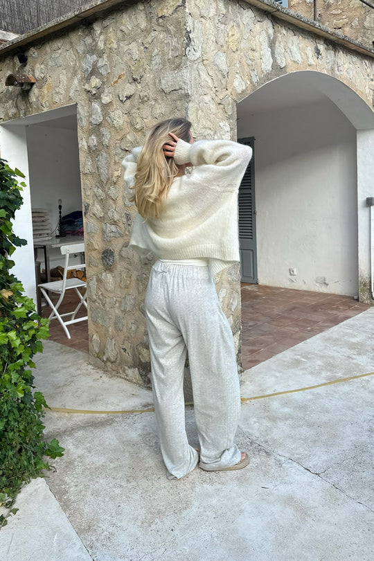Kvinde i afslappet, lys striktrøje og bukser står udendørs ved stenvæg. Komfortabelt tøj, afslappet stil, moderne mode.