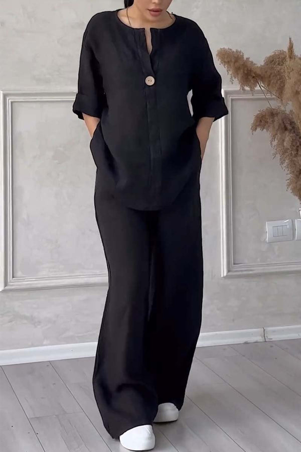 Kvinde iført sort loungewear-sæt med lange bukser og ærmer, stående på trægulv foran en lys væg med dekorative paneler.