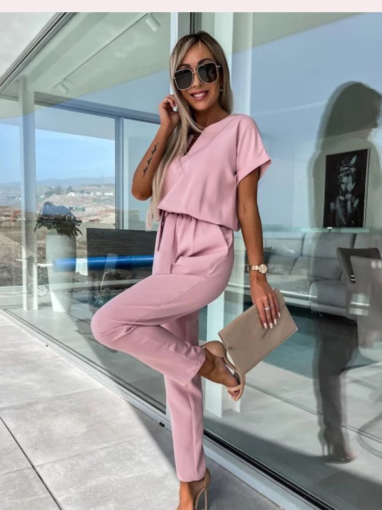 Kvinde i lyserødt jumpsuit står på moderne terrasse, holder beige clutch, iført solbriller. Stilfuldt sommeroutfit, mode, udendørs.