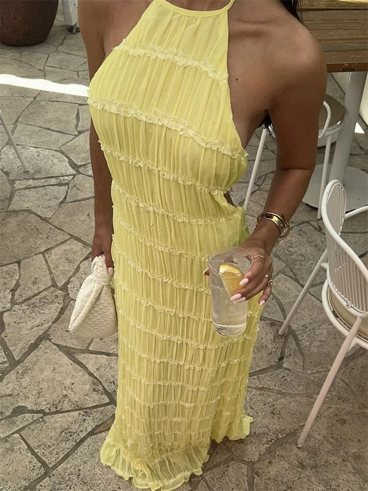 Kvinde i gul sommerkjole med halterneck, holder et glas vand med citron og en hvid taske. Perfekt til sommermode og udendørs begivenheder.