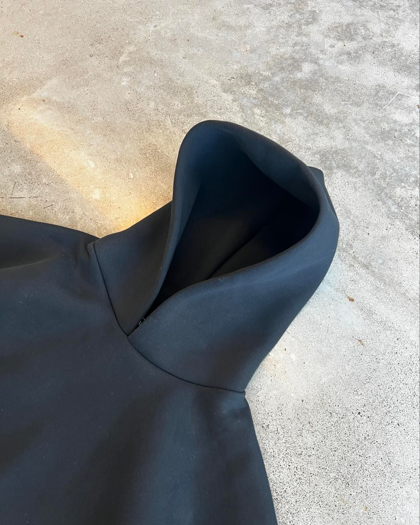 Sort asymmetrisk kjole med høj krave på grå betonbaggrund. Moderne design, minimalistisk stil, perfekt til modebevidste kvinder.