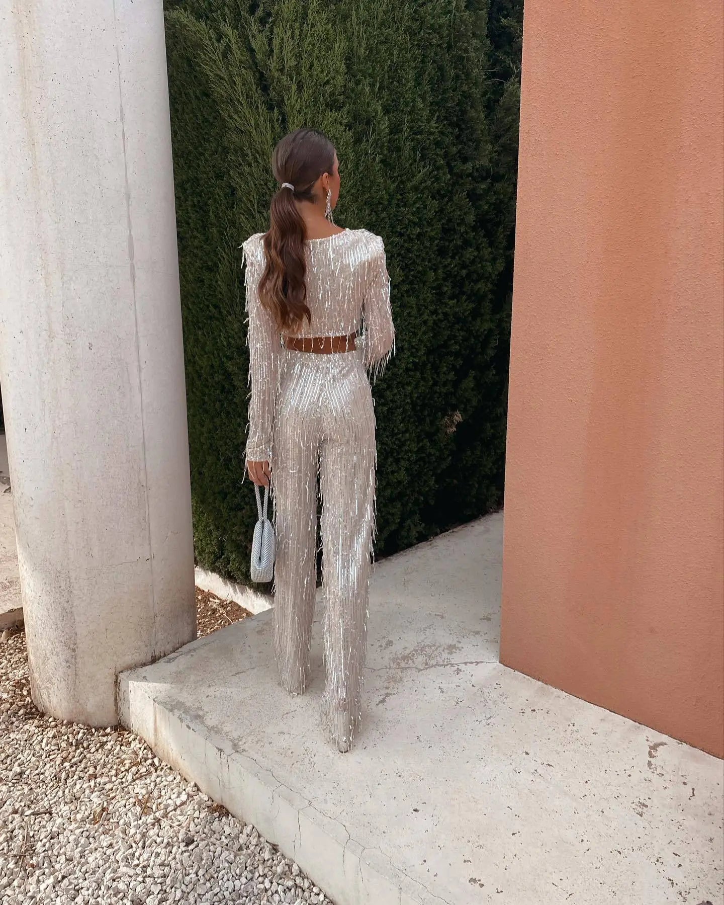 Kvinde i glitrende sølvdragt med langt hår i hestehale, står ved moderne arkitektur. Elegant mode, festtøj, udendørs fotoshoot.