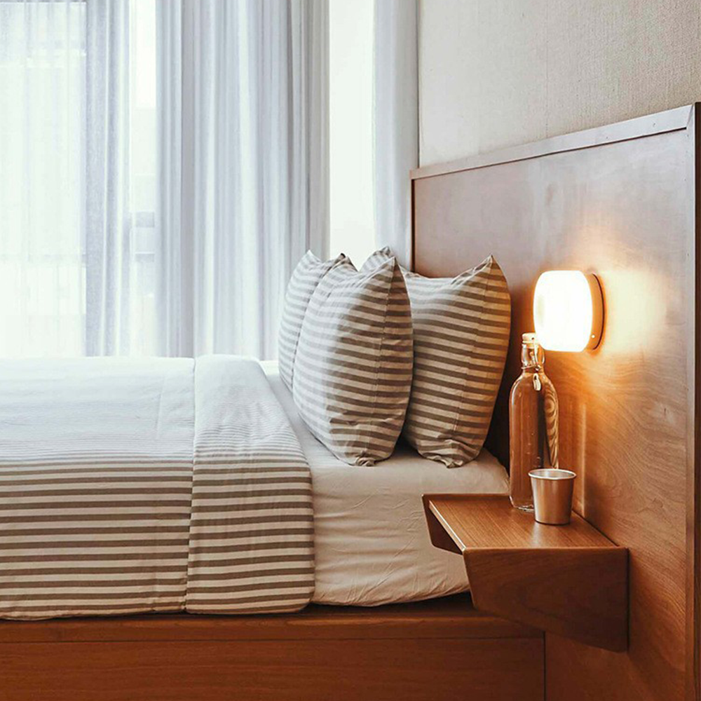 Moderne soveværelse med træramme seng, stribede puder, væglampe og natbord med glasflaske og kop. Lys og minimalistisk indretning.