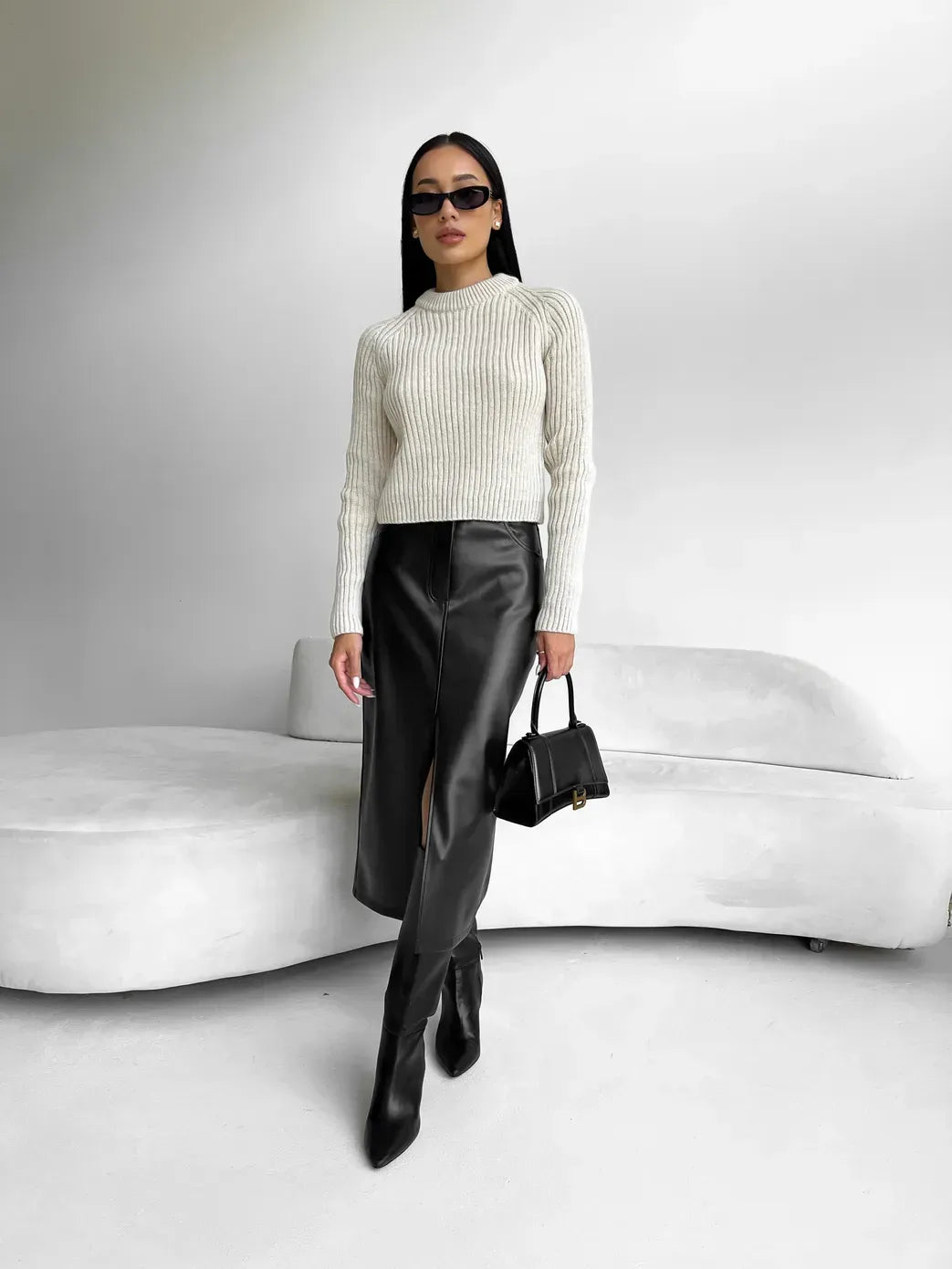Kvinde i moderne outfit: hvid striktrøje, sort lædernederdel med slids, sorte støvler og solbriller. Står foran hvid sofa, bærer sort taske.