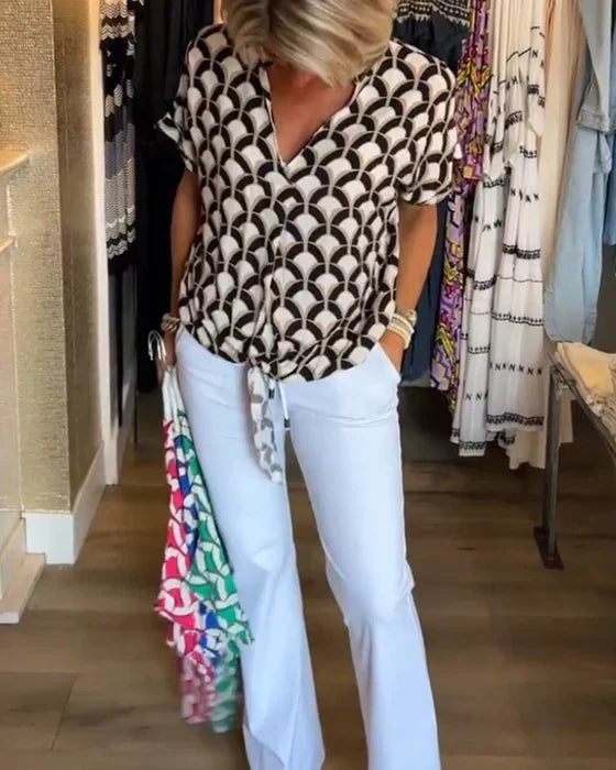 Kvinde iført mønstret bluse og hvide bukser i butik, holder farverige tøjstykker. Mode, shopping, stilfuldt outfit, moderne kvindetøj.