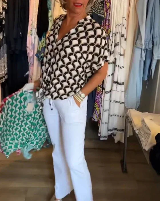 Kvinde i moderne tøjbutik iført sort-hvid mønstret bluse og hvide bukser, holder farverig skjorte. Mode, shopping, stilfuldt tøjvalg.