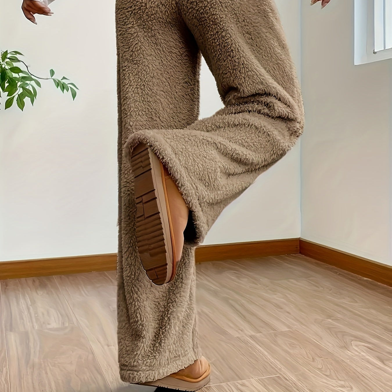 Brune fleecebukser, komfortabelt tøj, kvinde løfter benet, moderne indretning, trægulv, indendørs plante, afslappet stil, vintermode.