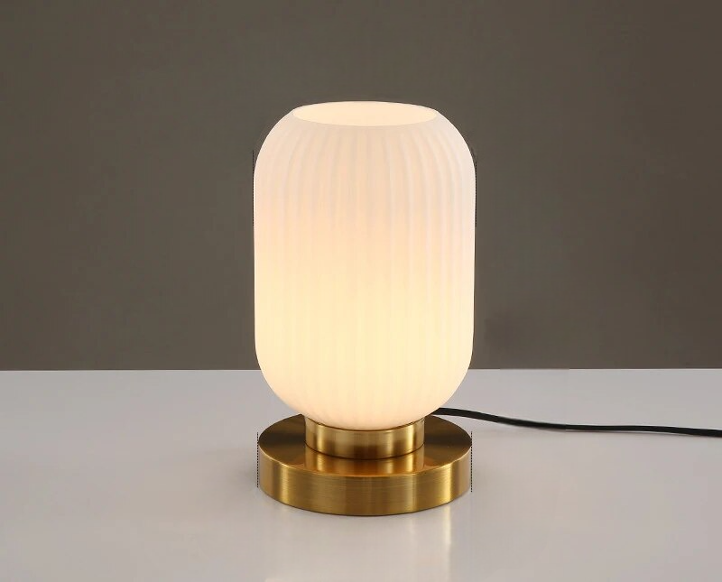 Moderne bordlampe med hvid, rillet glasskærm og gylden metalbase på hvidt bord. Stilfuld belysning til hjemmet, minimalistisk design.