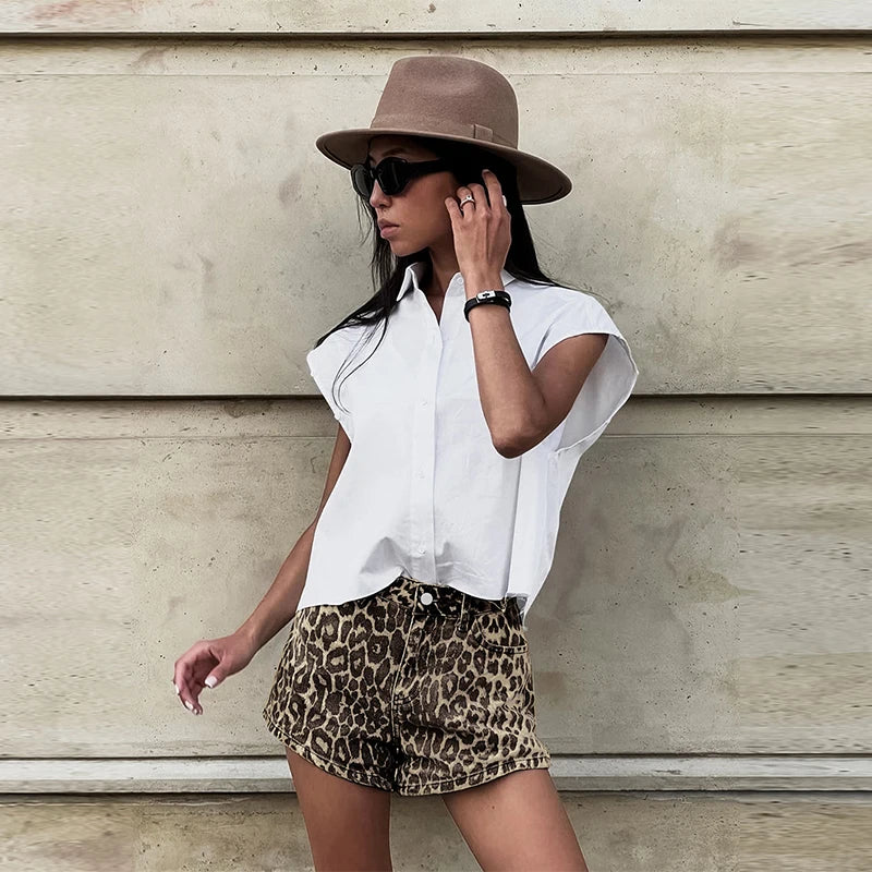 Kvinde i hvid skjorte og leopardmønstrede shorts, iført solbriller og brun hat, står op ad en beige væg. Mode, sommerstil, trendy outfit.