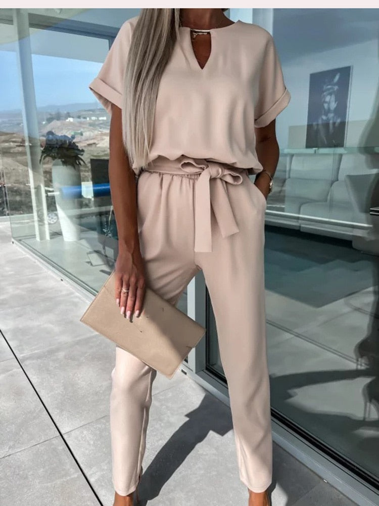 Kvinde i beige buksedragt med bælte, holder en matchende clutch. Står på terrasse med glasvægge. Elegant og moderne outfit til forår/sommer.