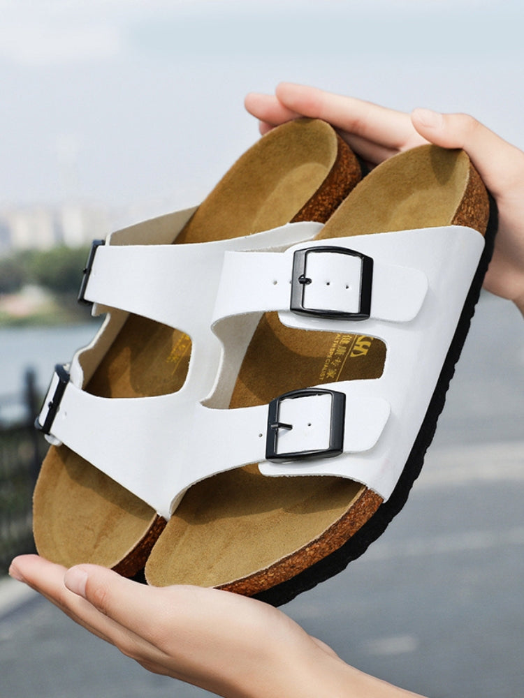 Hvide sandaler med dobbelt spænde, ergonomisk korksål, komfortabelt fodtøj til sommer, holdt i hænderne udendørs.
