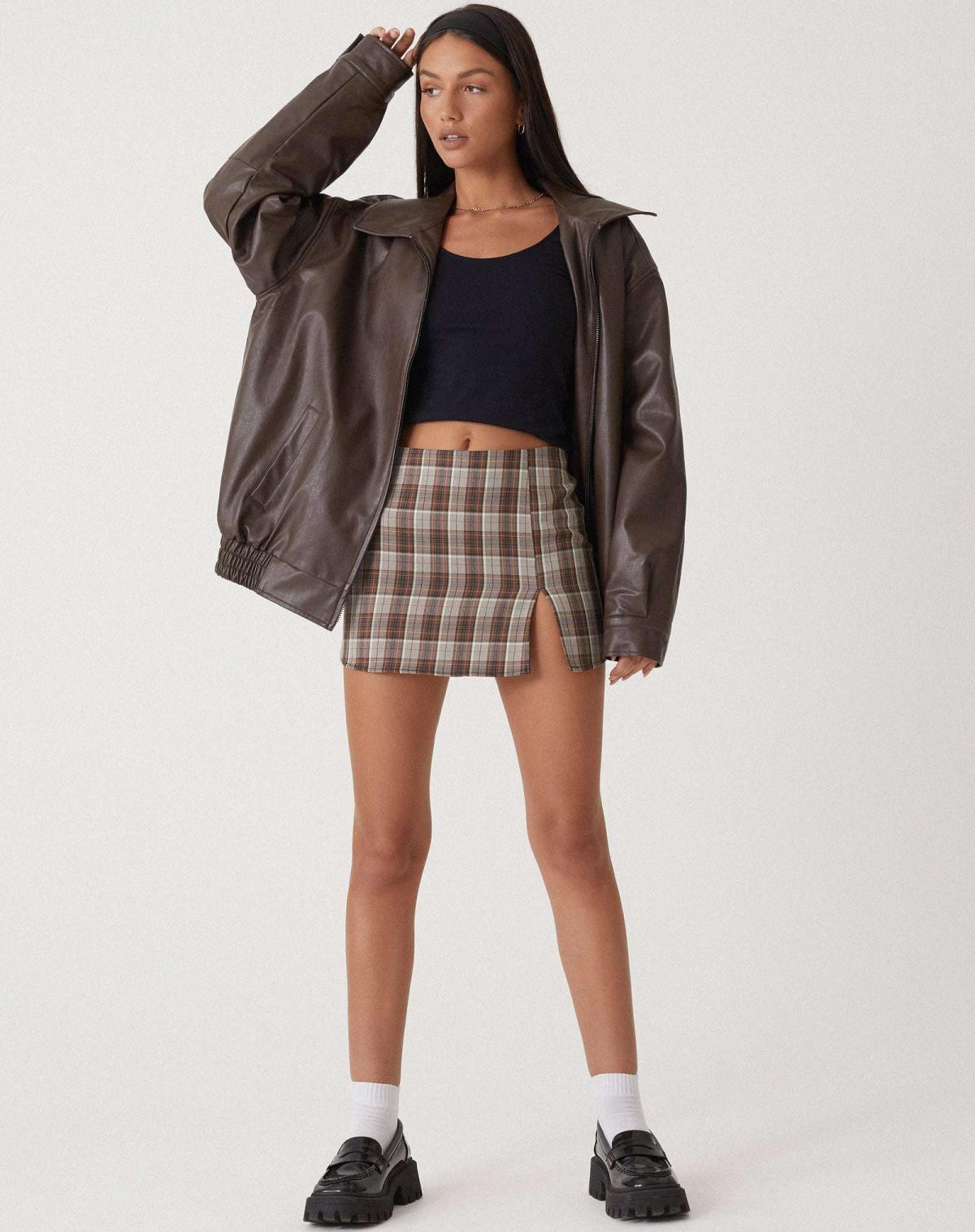 Kvinde iført brun læderjakke, sort crop top og ternet nederdel. Mode, streetwear, stilfuldt outfit, moderne tøj, ungdommelig look.