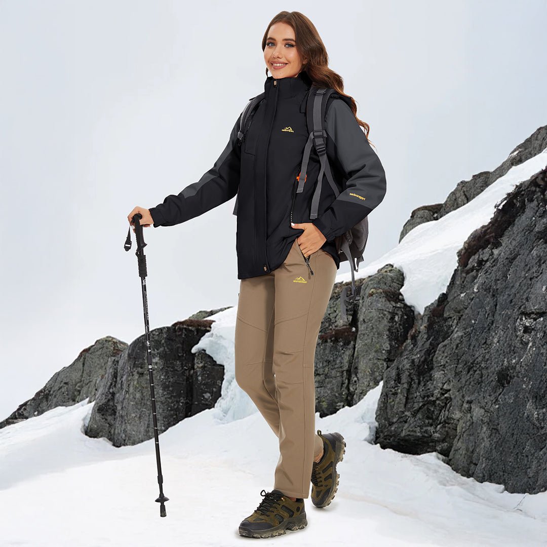 Kvinde iført sort vandrejakke og beige bukser, står på snedækket bjerg med vandrestav. Udendørs vintertøj, trekking, bjergvandring.