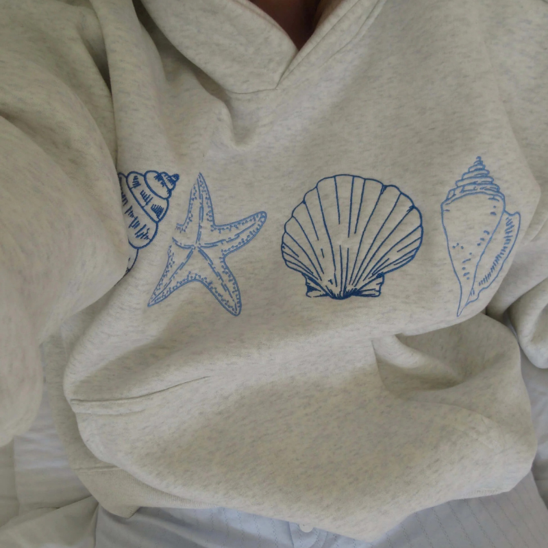 Grå sweatshirt med blå skaldyrsmotiver: konkylie, søstjerne, muslingeskal. Komfortabelt tøj, maritimt design, afslappet stil.