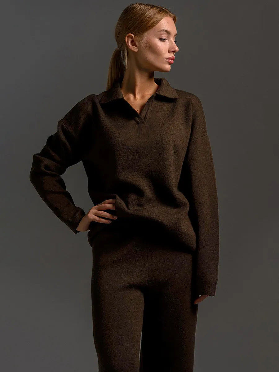 Kvinde iført mørkebrun sweater og bukser, poserer mod grå baggrund. Stilfuldt, minimalistisk outfit, efterårs- og vintermode, elegant look.