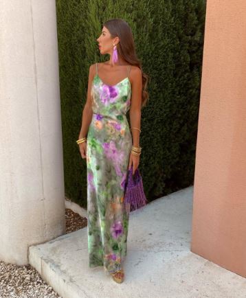 Kvinde i lang, blomsterprintet kjole med lilla og grønne nuancer, holder lilla frynsetaske. Står ved siden af grøn busk og orange væg.