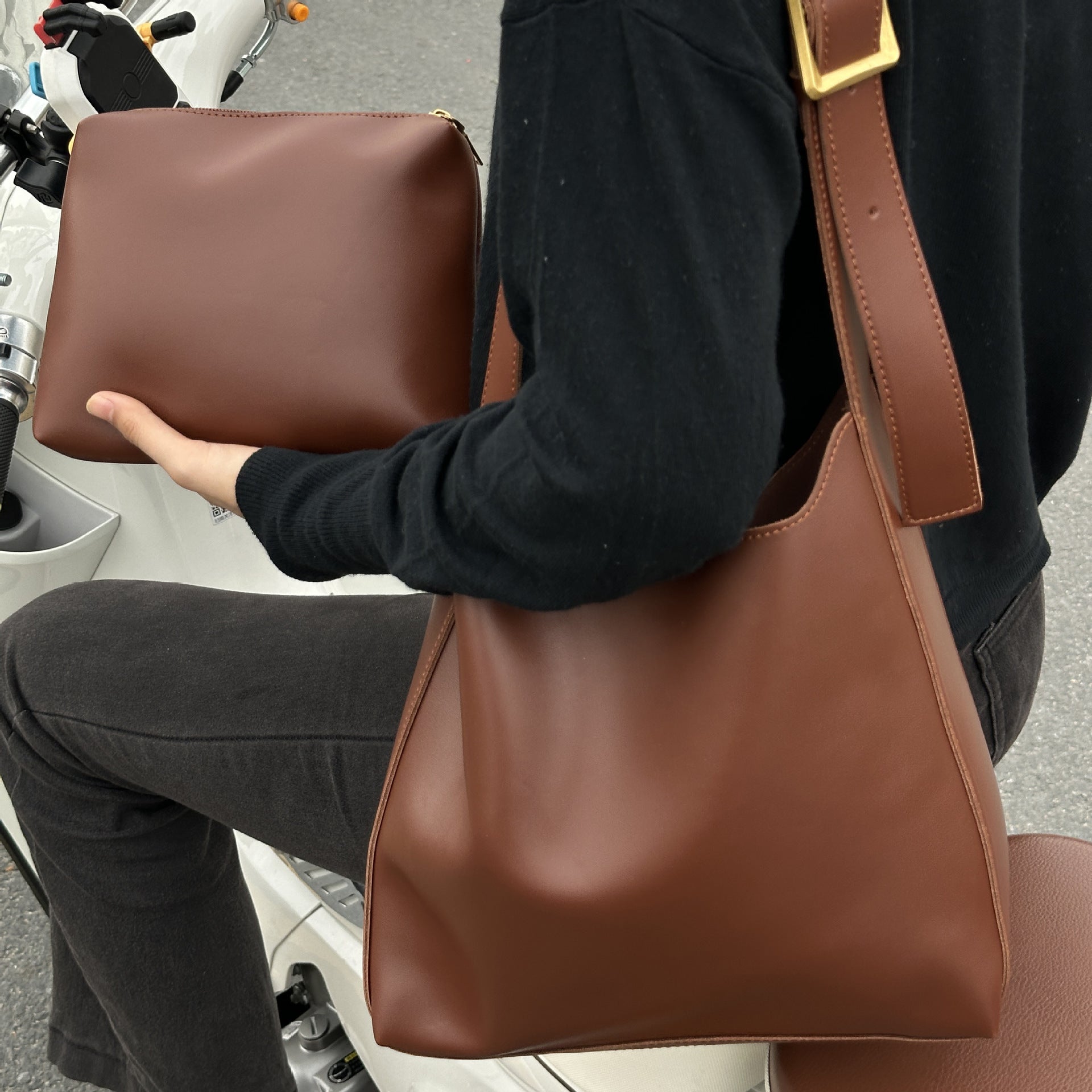 Brun læderhåndtaske og clutch i minimalistisk design, båret af person på scooter. Stilfuldt tilbehør til modebevidste kvinder.