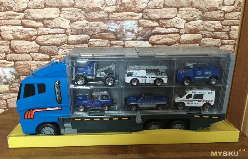 Legetøjslastbil med seks små politibiler i gennemsigtig opbevaringskasse, blå og hvid design, placeret på gul base foran murstensbaggrund.