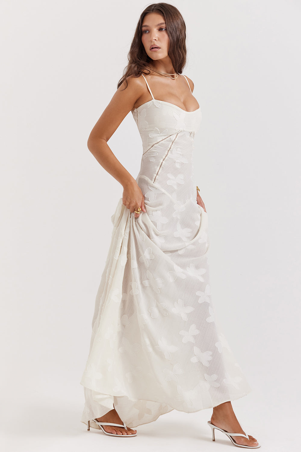 Kvinde i elegant, hvid sommerkjole med blomsterprint og tynde stropper. Perfekt til bryllup eller fest. Står i profil med hænderne i lommerne.