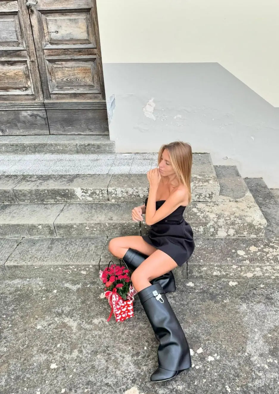Kvinde i sort kjole og høje sorte støvler sidder på stentrin med en buket røde roser. Elegant mode, udendørs, stilfuldt outfit.