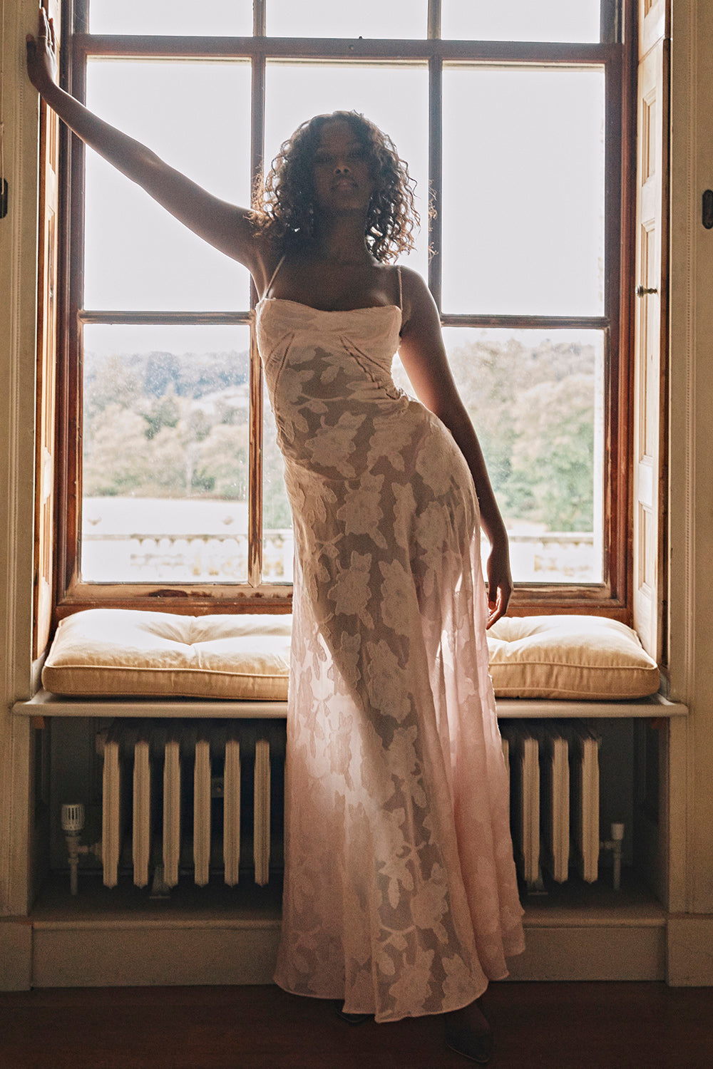 Kvinde i lang, lyserød kjole står ved vindue med udsigt til natur. Elegant mode, sommerkjole, indendørs, naturligt lys, stilfuld posering.