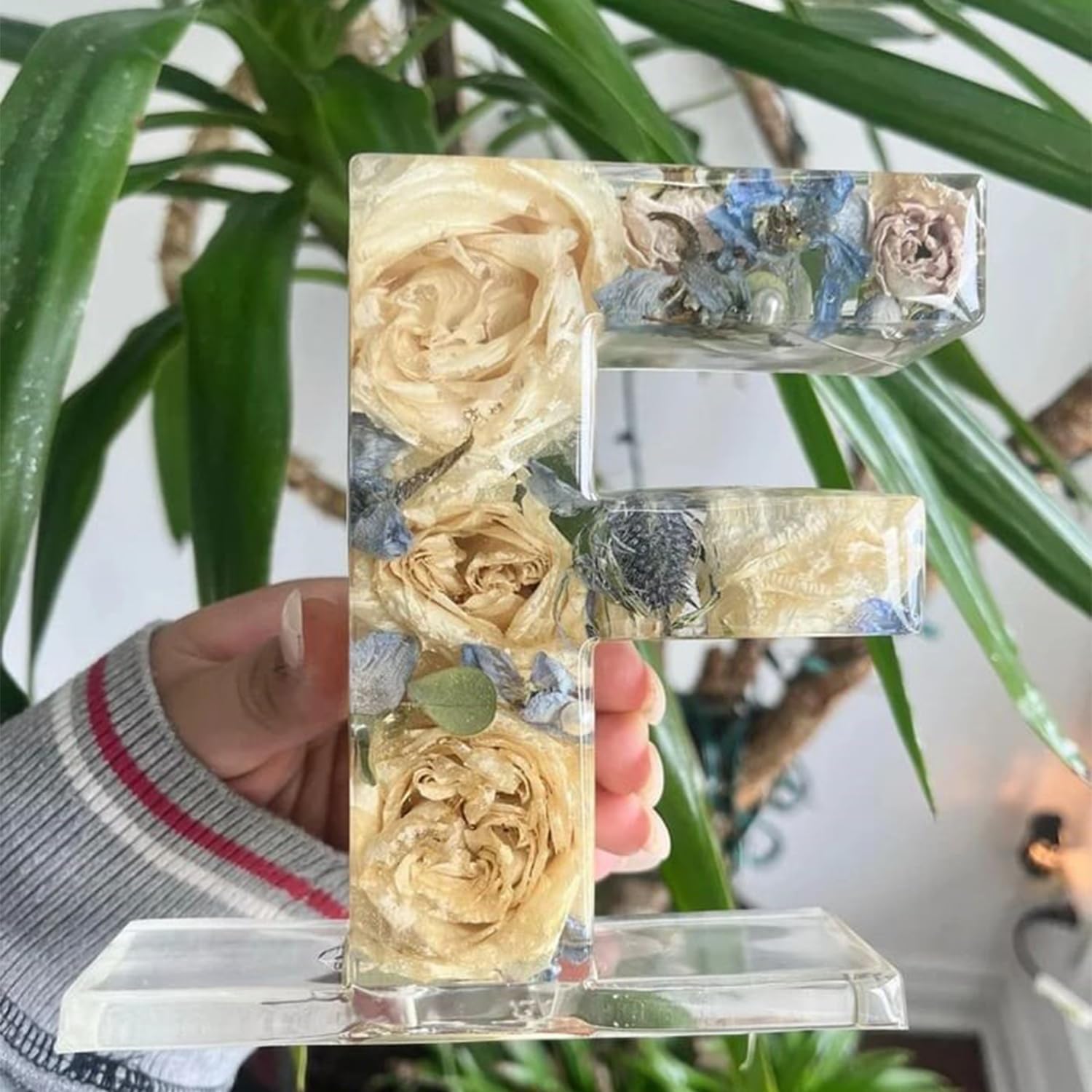 Klar harpiksbogstav "F" med indlejrede tørrede blomster, herunder roser og blå blomster, holdt foran en grøn plante. Dekorativ boligindretning.