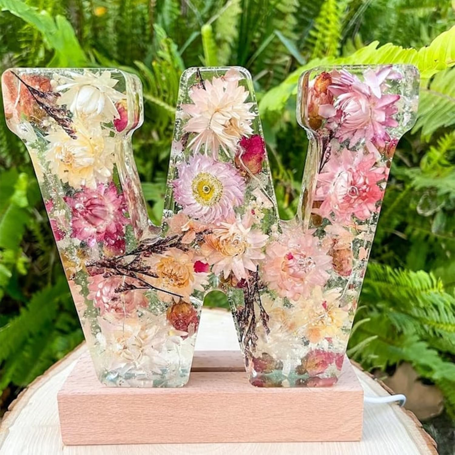 Dekorativt bogstav "W" i harpiks med indlejrede tørrede blomster, placeret på en træbase, omgivet af grønne planter. Perfekt til boligindretning.