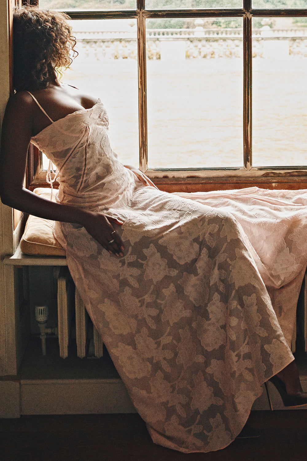 Kvinde i lyserød blondekjole sidder ved vindue, naturligt lys, elegant mode, romantisk stil, sommerkjole, afslappet atmosfære, indendørs portræt.