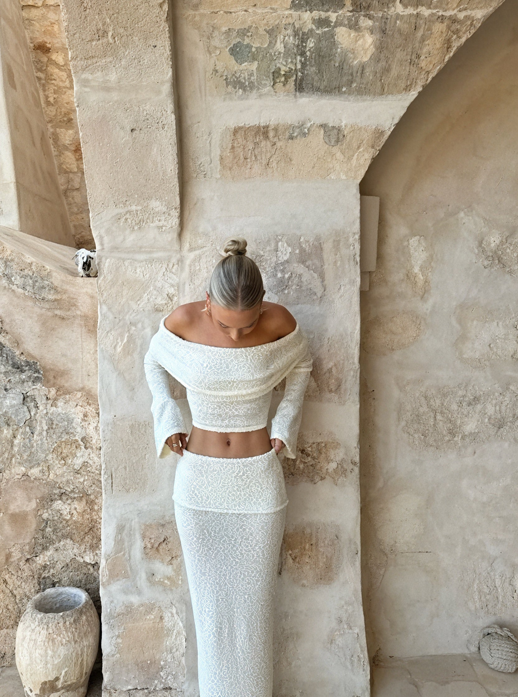 Kvinde i hvid, skulderfri kjole står mod rustik stenvæg. Elegant mode, minimalistisk design, moderne stil, skandinavisk æstetik.