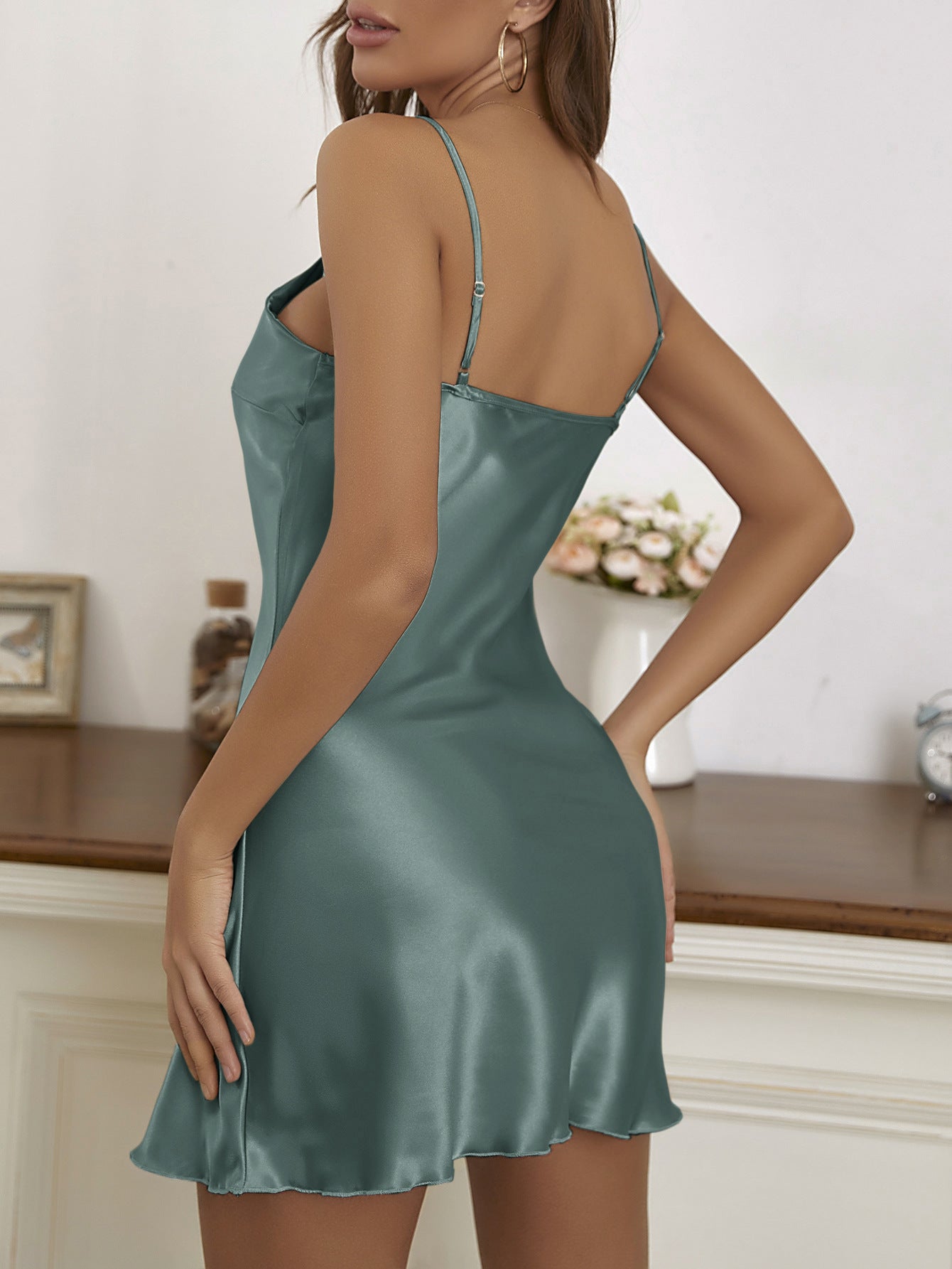 Kvinde i satin natkjole, grøn farve, set bagfra. Elegant design, tynde stropper, perfekt til aftenbrug. Moderne soveværelsesindretning i baggrunden.