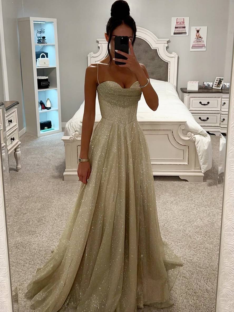 Kvinde i elegant, glitrende aftenkjole tager selfie i soveværelse. Kjolen har spaghetti-stropper og en fyldig nederdel. Luksuriøs mode, festkjole.