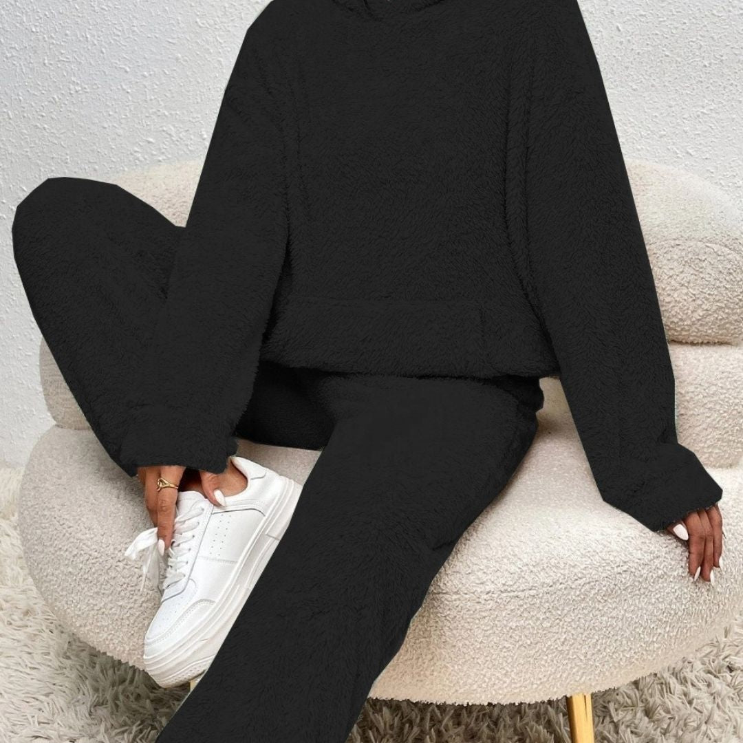 Kvinde iført sort fleece tracksuit sidder afslappet i en beige lænestol, iført hvide sneakers. Komfortabelt og stilfuldt loungewear til hjemmet.