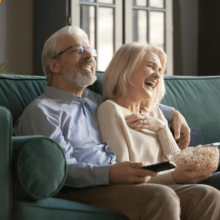 Ældre par sidder på grøn sofa, ser tv og griner, mens de deler en skål popcorn. Hyggelig hjemmeaften, afslapning, seniorlivsstil.