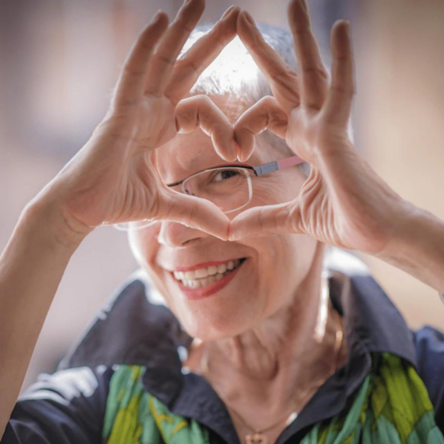Ældre kvinde med kort gråt hår og briller laver hjerteform med hænderne, smilende. Fokus på glæde og positivitet.